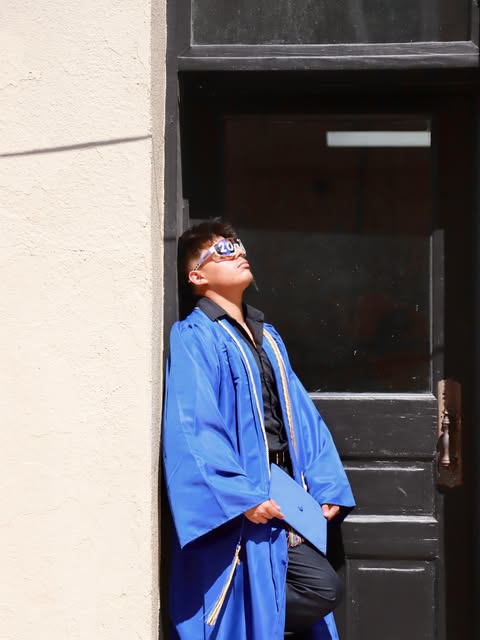 Graduate in a blue gown leaning in a doorway, cap in hand.